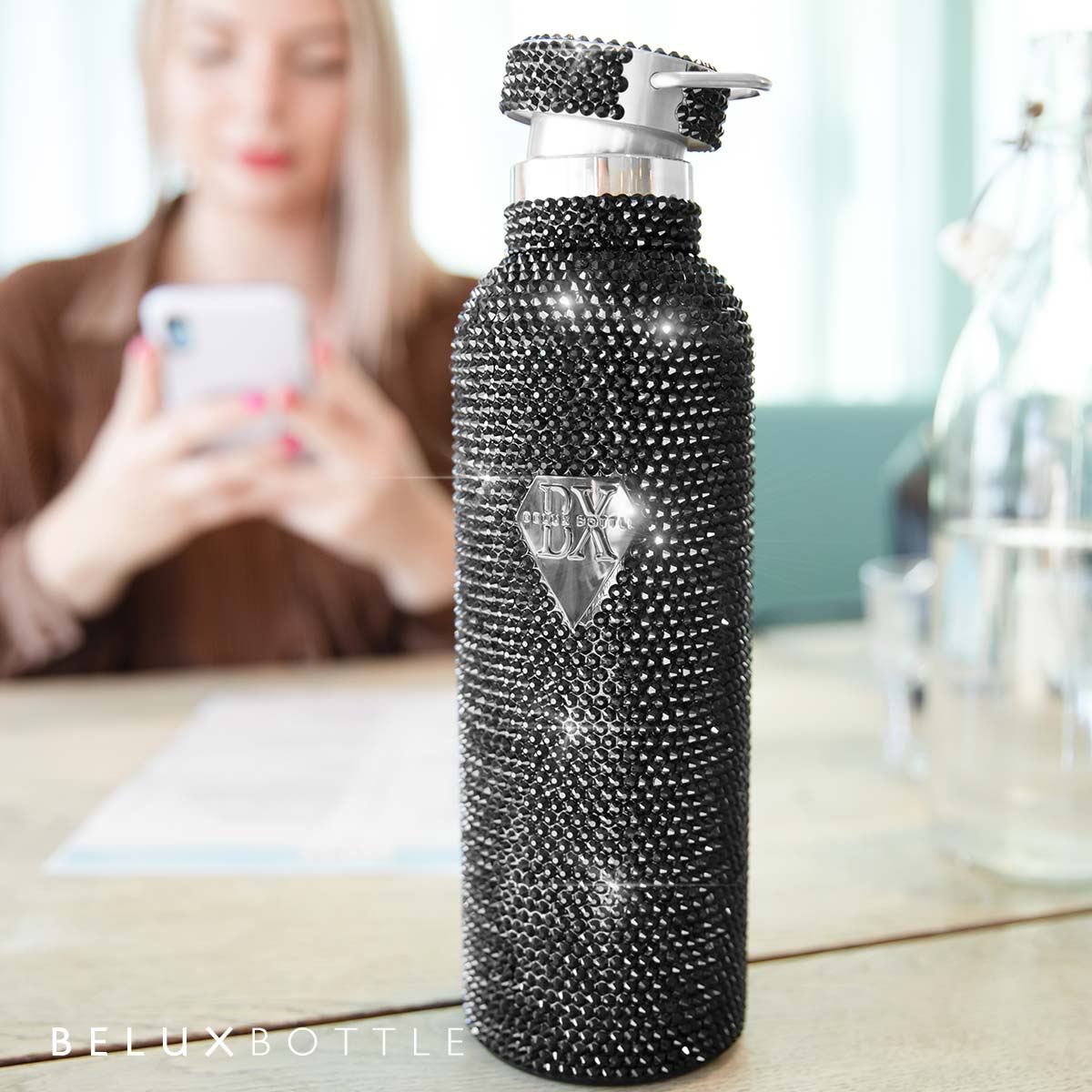 Stylish Belux Bottle Black Diamante Drink Bottle, with a woman engaged on her mobile in the background